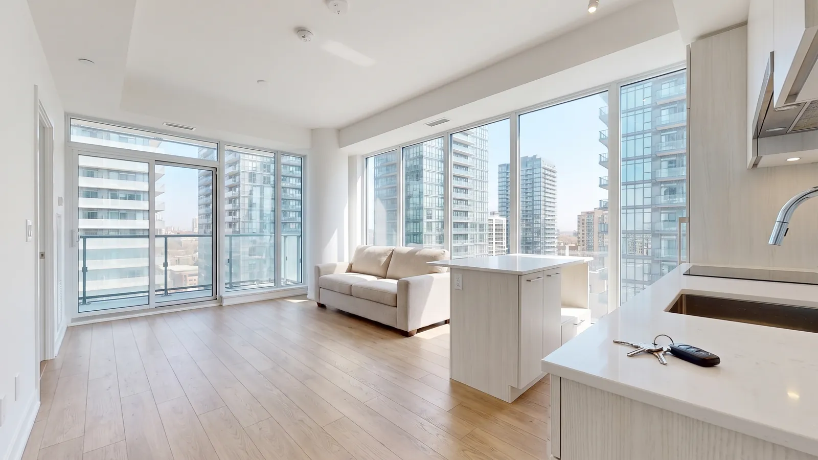 A bright, empty one-bedroom condo in Toronto with light oak floors, a beige sofa, floor-to-ceiling windows onto other glass towers and a set of keys on the kitchen counter