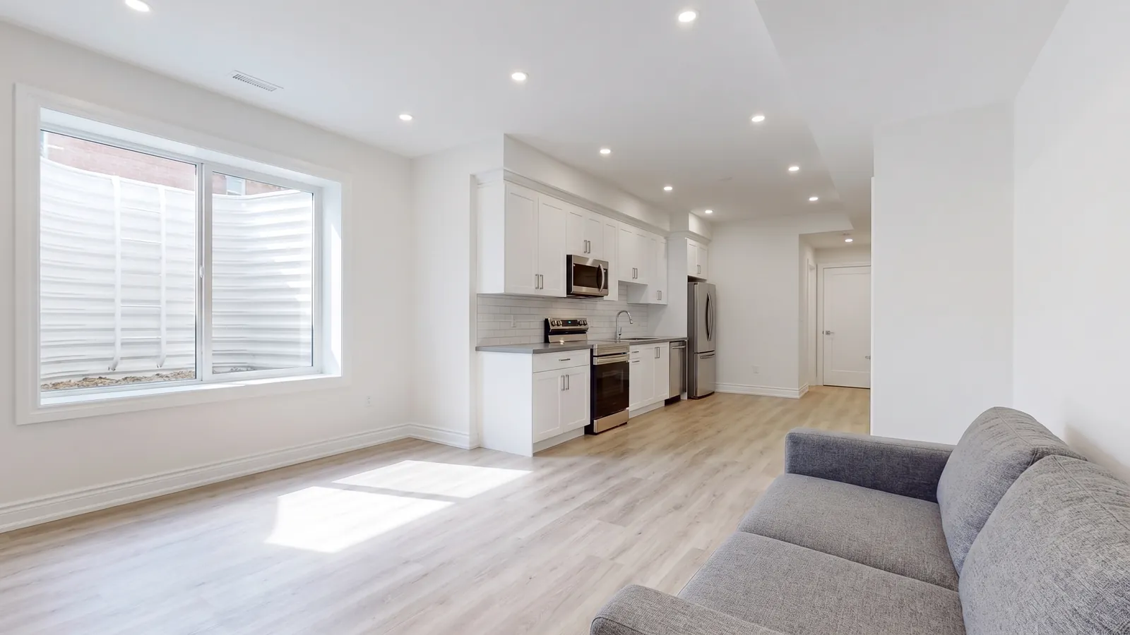 A bright, newly finished basement apartment with pot lights, light vinyl plank floors, a white kitchen, a grey sofa and a large window letting in daylight